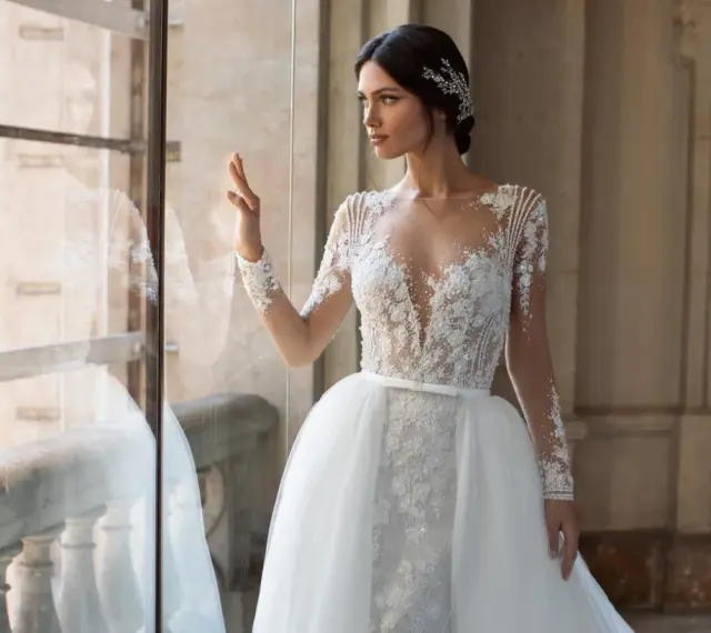 Panna młoda w sukni ślubnej z koronką i trenem, z ozdobną fryzurą w stylu bollywood, patrzy przez okno.