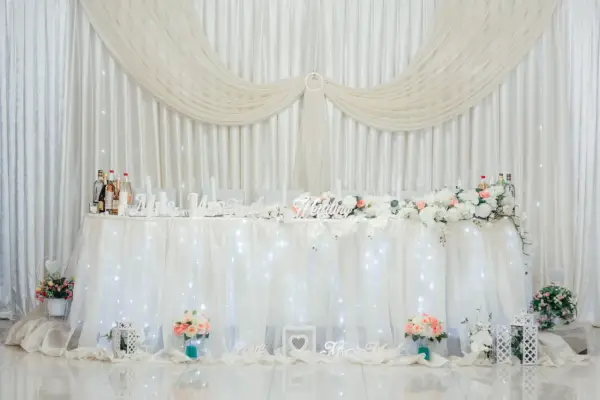 Elegancka ścianka za parą młodą inspiracje: białe zasłony, kwiaty, napisy "Wedding", "Love", "Mrs & Mr".